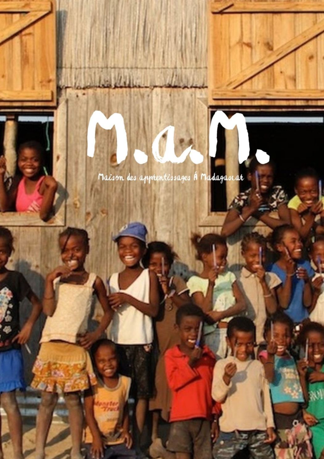 Des enfants d'Ampasifoty sourient devant une case en bois, sous le logo M.a.M.